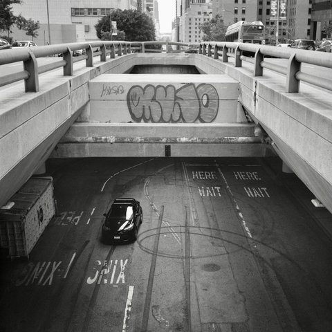 A black and white film photo. A vehicle is seen on the street below from the street stacked above it in downtown Los Angeles, California. Saturday, May 17, 2025.
