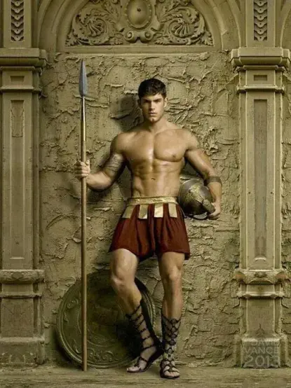 A photograph of a muscular young man with short, dark hair. He is dressed in a red kilt that reaches to his mid-thigh. Sandals reaching up two thirds to the knee decorate his legs. He holds a spear in his right and a helmet in his left. An aspis shield can be seen behind him in the background where it leans against a wall decorated with reliefs. He is standing between two columns, invoking Alektryon standing guard for Ares while he made love to Aphrodite.
