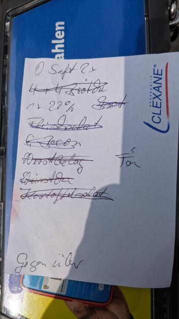 Bildbeschreibung von KI
Das Foto zeigt einen handgeschriebenen Einkaufszettel auf einem weißen Notizzettel mit dem Logo „CLEXANE®“ (ein Medikamentenname) am rechten Rand.

Der Zettel liegt auf einem blauen Untergrund, auf dem das Wort „Zahlen“ zu erkennen ist – vermutlich ein Kassen- oder Werbedisplay. Im unteren Bereich sieht man ein Smartphone mit geöffneter App, das von einer Hand gehalten wird. Der Einkaufszettel selbst ist von Sonnenlicht teilweise beschienen, was einen Schatten in der unteren rechten Ecke erzeugt.

Auf dem Zettel stehen mehrere handschriftliche Notizen, teils durchgestrichen – offenbar Dinge, die schon besorgt wurden. Hier ist der Inhalt in lesbarer Reihenfolge (teils schwer entzifferbar):

* O Saft 2x
* 4 x Rooter *(durchgestrichen)*
* * 20 % Brot *(durchgestrichen)*
* Schokolade *(durchgestrichen)*
* Speck *(durchgestrichen, eingekreist)*
* Ansteck... *(vermutlich „Anstecker“ oder „Ansteckzwiebeln“, durchgestrichen)*
* Winston *(durchgestrichen, wahrscheinlich Zigarettenmarke)*
* Kartoffelsalat *(durchgestrichen)*
* Ton *(rechts daneben notiert, evtl. „Tonic“ gemeint)*
* Gegenüber *(am unteren Rand handschriftlich notiert)*

Der Zettel wirkt wie eine spontane Einkaufsliste, teils mit humorvollen oder umgangssprachlichen Ergänzungen.
 