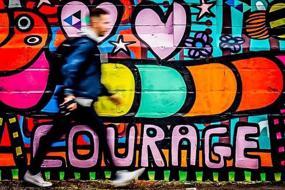 Graffiti. Eine bunte Raupe auf vielfarbigem Hintergrund mit rosa Herzen.
Schriftzug: COURAGE (französisch für Mut)
Ein junger Mann zielstrebig daran vorbei. So schnell, das seine Konturen verschwimmen
.