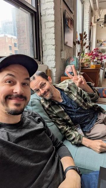 Rob and Kelly sit on a couch in our Toronto office, smiling for the camera. Kelly holds up a peace sign. Behind them is the clutter of the office, which includes flowers and a globe.
