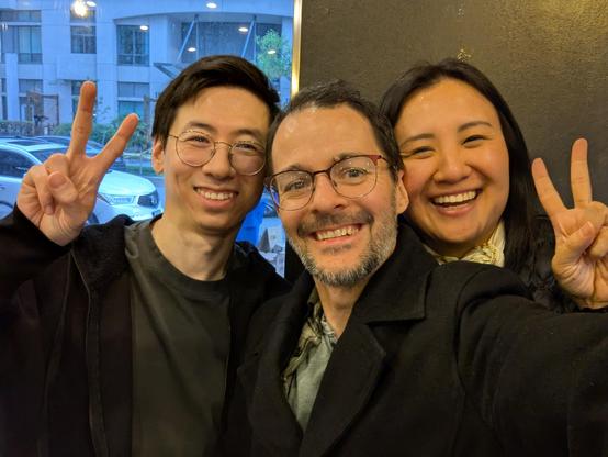   Jon, Kelly, and his spouse hold up peace signs while smiling for a selfie. 
