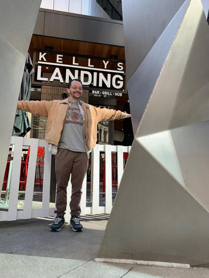 Kelly smiles for the camera with his arms outstretched while standing between two geometric, metal pillars.  The white fence behind him demarcates a patio area for the restaurant behind Kelly. It's called "Kellys Landing."

