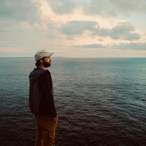 Colin Manson in a cap and casual clothes stands on a cliff, gazing at the ocean under a cloudy sky. Calm and contemplative atmosphere. 