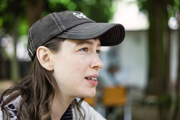 Portrait photo of a woman with long hair wearing a cap. Blurry trees and a grey wall in the background.