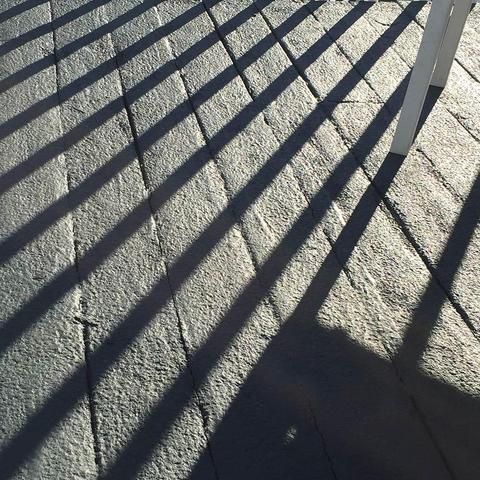 Diagonal shadows cast by railings create a geometric pattern of dark parallel lines across a tporch floor. The shadows alternate with bright sunlit strips, forming a striking linear composition.