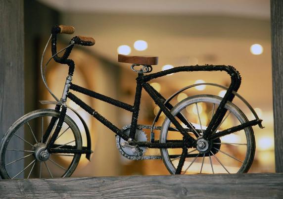 The image features a decorative bicycle model, intricately crafted with a black beaded frame. The bicycle is positioned on a wooden surface, with its wheels, handlebars, and seat clearly visible. The frame is adorned with black beads, giving it a textured appearance, while the wheels are made of metal with spokes. The handlebars have wooden grips, and the seat is also made of wood, adding a touch of natural texture. The background is softly blurred, with warm, bokeh lights suggesting an indoor …