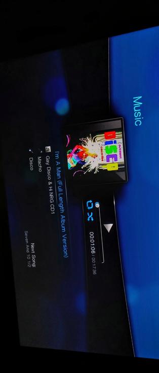 The photograph composed in total darkness shows a screen displaying a music player interface. The background is dark with a blue section on the right side labeled "Music." The screen displays the title "I'm A Man (Full Length Album Version)" by Gay Disco & Hi NRG, with an album cover featuring colorful text spelling "DISCI" and a vibrant image. The track is from the "Disco Macho" collection. The playback time is shown as 00:01:06 out of 00:17:36. The next song is "Seven And 10 1/2." The interface includes playback controls such as a rewind and fast-forward button, and a progress bar.