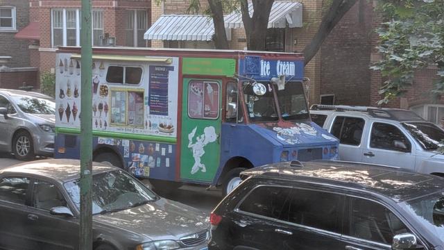 Photo of an ice cream truck on a residential street on the NW side of Chicago. The truck has a blue front with a mural on the hood (can't see it clearly), and what looks like a silhouette of a chef on the side door, which is painted green.