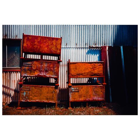 Four rusted metal containers are stacked in pairs against a corrugated sheet metal wall. Each bin wears a thick coat of oxidized orange and red, flaking like they’ve spent the last decade weathering typhoons and neglect. The building’s siding, a faded beige and silver, adds to the scene’s industrial fatigue.