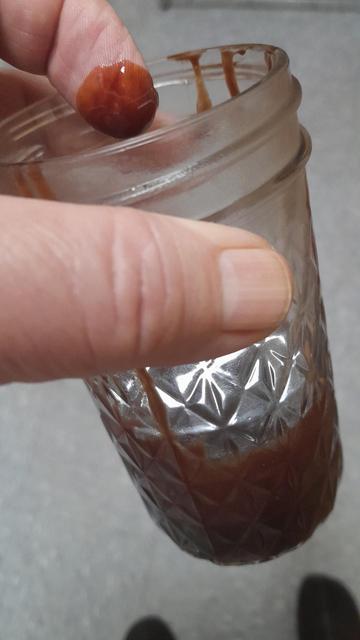 A glass with some chocolate syrup at the bottom and a hand holding it at the top with the index finger covered in chocolate syrup
