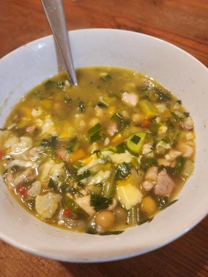White bowl of soup with a spoon in it. The soup is golden coloured and thick with diced, multicoloured vegetables.