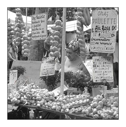 Marktfrau an ihrem Marktstand zwischen Knoblauch und Zwiebelketten. Tafeln mit französischen Informationen und Preisen zu den Produkten. 