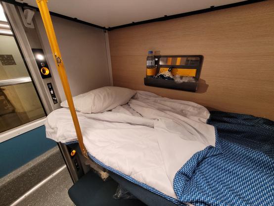 Zoom on a couchette, showing an open blanket with integrated sheet, a power plug and light in each bed, and a small shelf with a provided water bottle.