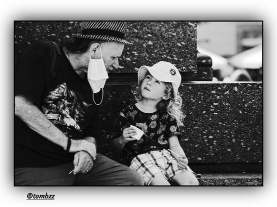 An elderly man and a young girl are sitting together on the steps of a monument. The girl is wearing a t-shirt with a cat print, checkered shorts, and a baseball cap with a small emblem on it. The man is dressed in a Harley Davidson t-shirt featuring a large wolf print, pants, and a striped hat. Their relationship appears to be close, as they sit together and engage in conversation. The photograph is in black and white.