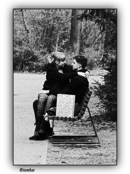 The photo depicts a couple in a park. The young man is sitting on a bench, while the young woman is on his lap, their closeness indicating a romantic bond. She is wearing fishnet stockings, a short skirt, and chunky platform shoes, with her long hair cascading freely over her shoulders. The young man is dressed in dark pants, a t-shirt, and an open jacket. A paper gift bag sits on the bench beside them, suggesting a special occasion. In the background, trees and bushes add to the intimate atmosphere. The photograph is in black and white, emphasizing its nostalgic and emotional tone.