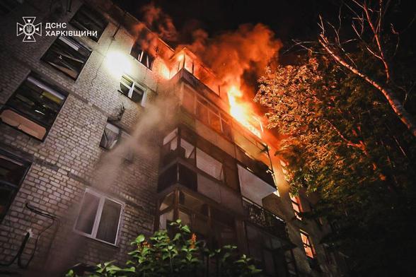 The image shows a nighttime scene of a building on fire. The fire is concentrated on the upper floors of the structure, with bright orange flames and thick black smoke billowing out of the windows. The building appears to be a multi-story residential or office complex. The fire is intense, and the heat can be felt through the image. In the foreground, there are trees and some greenery, adding contrast to the dark and fiery background. The photo is taken from a low angle, looking up at the building, emphasizing the height and severity of the fire. There is a logo in the top left corner, which seems to belong to an emergency service, possibly indicating that firefighting efforts are underway.