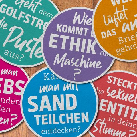 Verschiedene farbige Bierdeckel auf einem Holztisch ausgebreitet. Darauf stehen Forschungsfragen, z.B. "Wie kommt die Ethik in die Maschine?" Photo: DESY