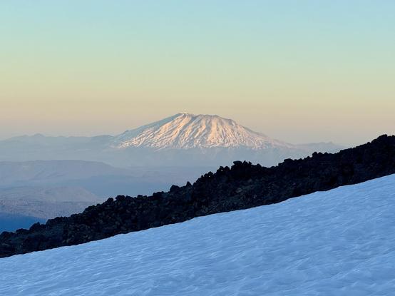 A view of Mount Saint Helens at sunrise from the side of Mount Adams. The snow covered volcano has a flattish top after erupting in 1980. A white snow field and dark rocky ridge are in the foreground. The skies shade from pinks to yellows to greens.