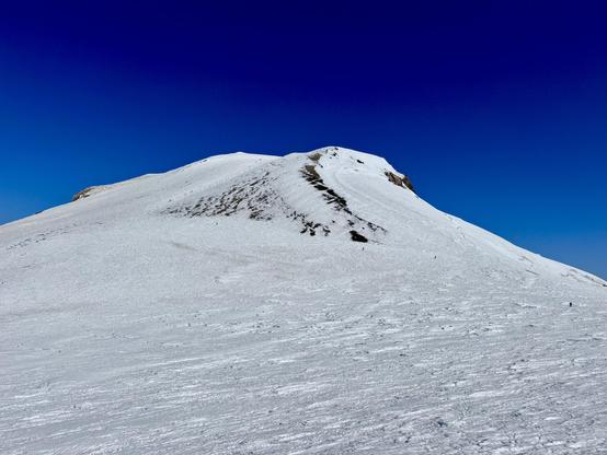Mountaineers zigzag across and up the final slopes to reach the true summit of Mount Adams. Most of the volcano is covered in snow at this elevation. The smell of sulfur fills the air at times. Dark blue skies.