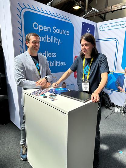 The pictures shows the Drupal booth in the background and Fredrick Wouters and James Abrahams standing at the podium in front of it.