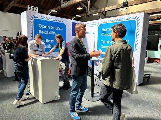 The picture shows the Drupal booth in the background and a few members in front of it engaged in small group discussions.