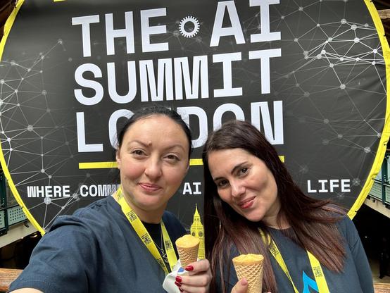 The picture is a selfie of Lenny Moskalyk and Catia Penas in front of The AI Summit London poster holding ice cream cones looking at the camera.