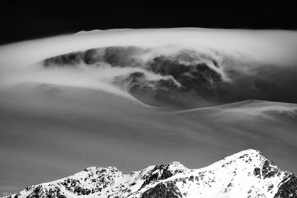A black and white photograph of cloud formations swirling above rugged mountain peaks in the Lofoten Islands, Norway. The mountains are capped with snow, their outlines standing in contrast to the smooth, shifting patterns in the sky. The scene captures a moment of stillness in the landscape below, set against the movement and drama of the weather above.