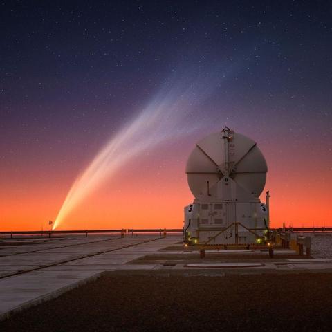 A sunset sky with orange tones at the bottom and a starry night above. In the lower left, just above the horizon, is a comet whose massive tail stretches up to the upper right, arcing over a radio telescope at ESO's Paranal Observatory in Chile.