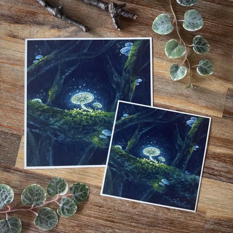 A photo of two square prints, one large, one small, placed on a wooden table and surrounded by leaves and branches for decoration. Both prints feature the same painting: White glowing mushrooms on mossy branches high up in the trees of a dark forest.
