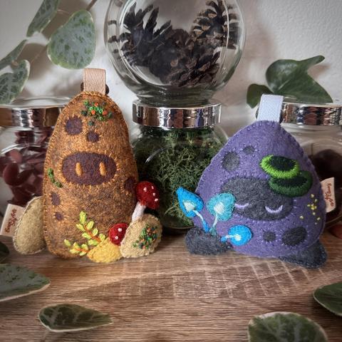 A photo of two felt bag charms in the shape of little rocky forest spirits. The left one features brown tones, amanita mushrooms, fern and moss embroidery. The right one features purple tones, glowing blue mushrooms, embroidered lichen cups and tiny yellow fungus spores. In the background glass jars with moss, dried flowers, pine cones, and acorns are visible.