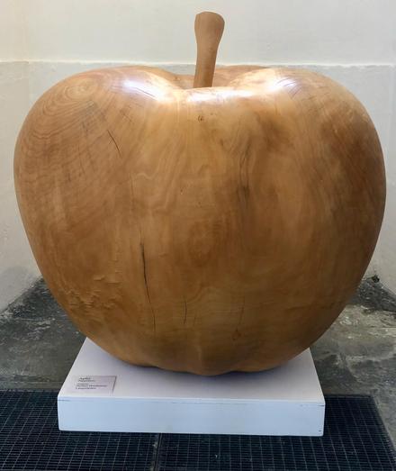 Eine Skulptur aus Holz. Ein Apfel, der ganz naturgetreu und etwas 80cm hoch ist. Steht auf einem weißen Sockel vor weiß getünchter Wand. Farbe des Apfels ist naturbelassenes Holz.