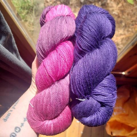 My hand, holding two skeins of yarn - pink and pinky purple on the left and a rich purple on the right.