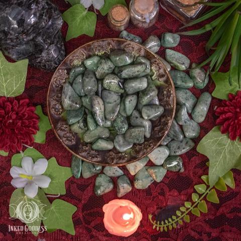 Several specimens of the "Grossularite Tumbled Gemstone" product are arranged on a dark lace-covered surface. Several more specimens fill a metal bowl. They are about than 1 inch across, tumbled smooth and highly polished. Presented by Inked Goddess Creations.