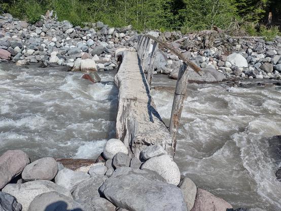 A "bridge" crossing a turbulent glacial river made of a log log spilt in half, with a few bits added at the end, set into the rocks on either end and a handrail on one side made of some upright sticks holding up long smaller sticks between them.