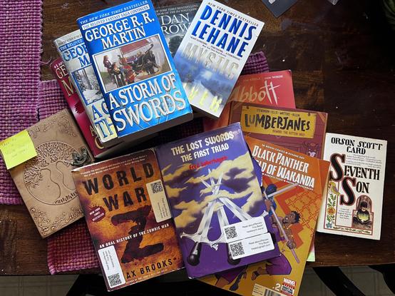 Stack of books with QR label stickers
A Game of Thrones - George R. R. Martin.
A Storm Of Swords - George R. R. Martin.
A Feast For Crows - George R. R. Martin.
Mystic River - Dennis Lehane
Angels And Demons - Dennis Lehane
World War Z - Max Brooks
Seventh Son - Orson Scott card
The Lost Swords: The First Trio - Fred Saberhagen
Lumberjanes Vol 1 - ND Stevenson
Black Panther: World Of Wakanda
Abbot (free comic day edition) - Saladin Ahmed
And a leather bound blank journal with carving on cover w…