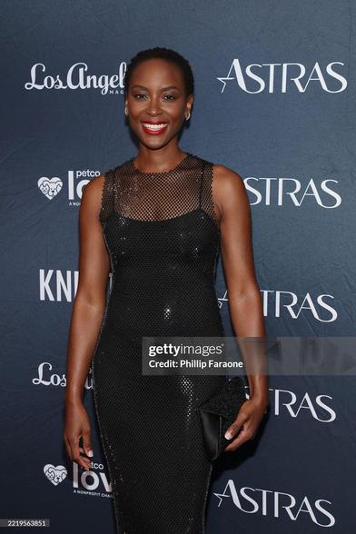 Tracy Ifeachor smiling on the red carpet of the Astra TV Awards. She wears a black form-fitting net dress without sleeves + a strong red lip.
