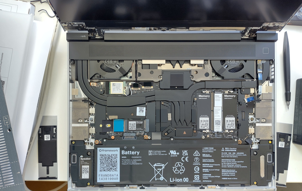 **TL;DR:** This is the internal view of a Framework Laptop showing modular components like battery, memory, storage, fans, and speakers.

**Text Extracted:**

* framework
* Battery
* 65Wh
* FRANDBATA
* This battery is replaceable
* Memory (DDR5 Channel 0 & 1)
* Storage
* Speaker
* Mainboard
* Li-ion 00
* Rechargeable Li-ion Battery
* Various certification logos (UL, CE, UKCA, etc.)

**Image Description:**
This photo shows the inside of an open Framework Laptop, emphasizing its modular and repairable design. At the bottom is a large rectangular battery labeled “framework” with a QR code. Above that are two memory modules (DDR5) and an M.2 SSD labeled "Storage." Two cooling fans are placed on the upper left and right sides. There's a speaker on the bottom left, visible wiring, and several QR codes for component identification. A pen and removed part are seen to the side, indicating active assembly or upgrade.
