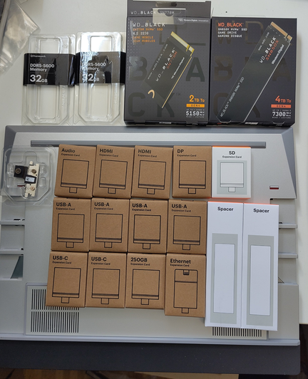 **TL;DR:** Collection of Framework Laptop components including memory, SSDs, expansion cards (USB, HDMI, etc.), and spacers, laid out on a device chassis.

**Text Extracted:**

* DDR5-5600 Memory 32GB (x2)
* WD\_BLACK SN770M SSD 2TB
* WD\_BLACK SN850X SSD 4TB
* Audio Expansion Card
* HDMI Expansion Card (x2)
* DP Expansion Card
* SD Expansion Card
* USB-A Expansion Card (x4)
* USB-C Expansion Card (x2)
* 250GB Expansion Card
* Ethernet Expansion Card
* Spacer (x2)

**Image Description:**
The image displays various modular components for a Framework Laptop arranged on top of a gray laptop chassis. At the top are two sticks of 32GB DDR5 RAM and two WD\_BLACK NVMe SSDs (2TB and 4TB). Below them are numerous cardboard boxes, each labeled with the type of expansion card inside, such as USB-A, USB-C, HDMI, DisplayPort (DP), Ethernet, SD card, 250GB storage, and Audio. There are also two white spacers and a small camera module. The layout shows the system’s high modularity and user-customizability.
