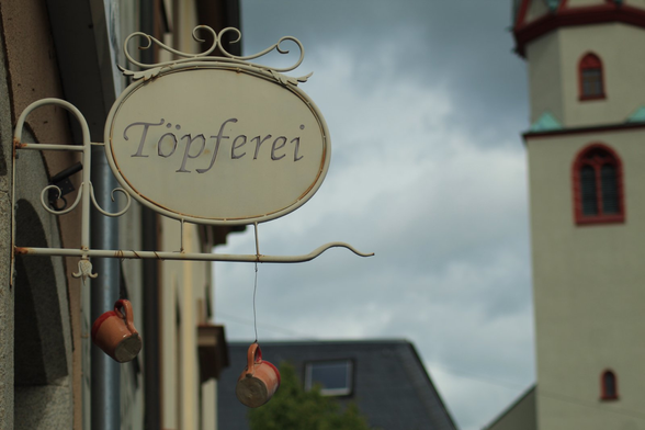 Nahaufnahme eines Ladenschildes, auf dem "Töpferei" steht. Zwei Krüge hängen daran herunter. Im Hintergrund ein Kirchturm im Anschnitt.