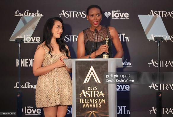 Tracy Ifeachor and Arun Storrs at the Astra TV Awards, accepting te prize for "The Pitt". Tracy wears a black form-fitting net dress without sleeves + a strong red lip. Arun wears a mini-dress in gold patterned fabric.