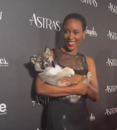 Tracy Ifeachor on the red carpet of the Astra TV Awards. She is holding a puppy in her arms and is smiling. Tracy wears a black form-fitting net dress without sleeves + a strong red lip.
