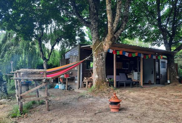 under a large tree a tagine cooks on the ground beside a colourful hammock and rustic barbecue in front of a wooden shack