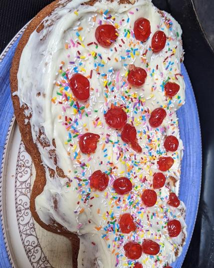 A white cake with sprinkles and cherries on top vaguely in the shape of raggedy ann jumping rope.
