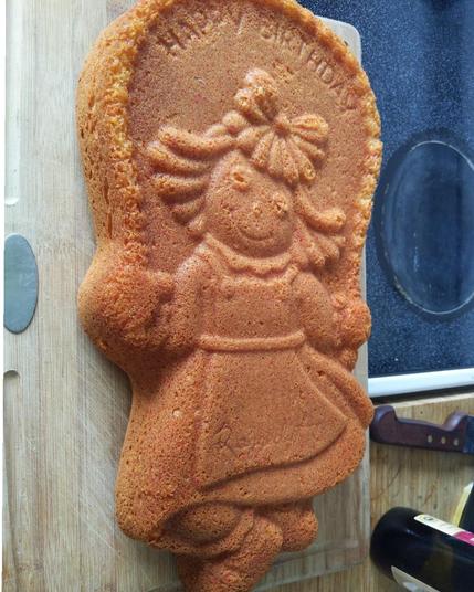 An un-iced cake showing an image of Raggedy Ann skipping with a jump rope. (Raggedy Ann looks like a little girl with a bow in her hair wearing a dress with an apron)