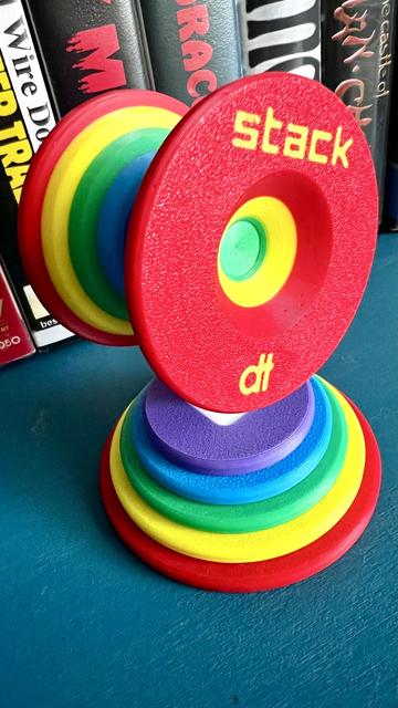 A rainbow 3D printed Stack yo-yo, by Dale Tubat, atop its rainbow 3D printed stand. Spines from various horror vhs peek from behind. 