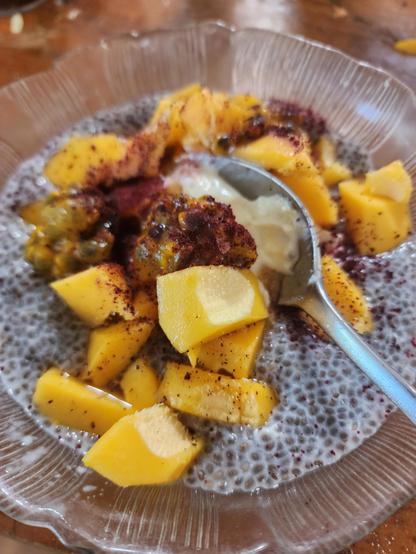 A glass breakfast bowl with spoon in it, filled with fruit   yoghurt, chia porridge, with dark red sprinkles on top.