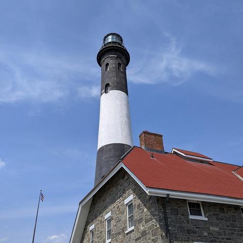 A tall lighthouse with distinctive black and white horizontal stripes stands against a blue sky with white clouds. The lighthouse features a black lantern room at the top with a glass enclosure. At its base is a stone keeper's house with gray stone walls, white-trimmed windows, and a red clay tile roof. An American flag flies on a flagpole to the left of the structure.