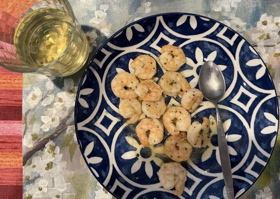 A serving of pink sauteed shrimp in a big wide bowl with a strong blue and white graphic design. A long iced tea spoon sits in the bowl too. A checkerboard faceted squat water glass sits beside it, half filled with white wine. Both sit on a watercolor pale green white and brown cherry blossom placemat. A strongly colored striped tablecloth in shades of hot pink and orange can be seen in one corner below the placemat