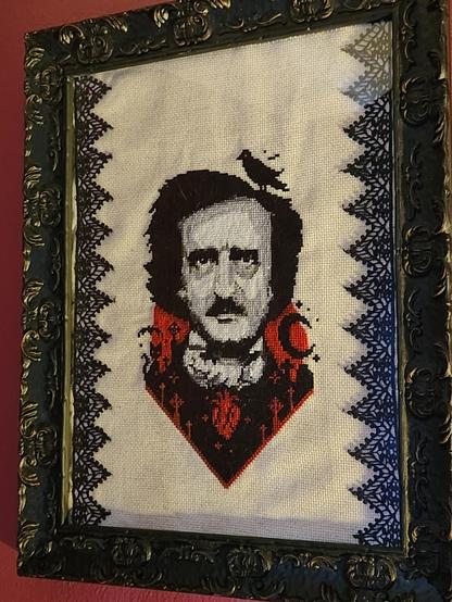 A counted cross-stitch portrait of Edgar Allan Poe in an ornate Victorian-style black frame on a magenta wall